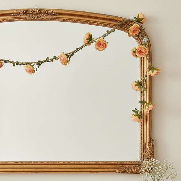 Decorative gold-framed mirror with floral garland on a light wall, books, and a vase with flowers on a surface below.