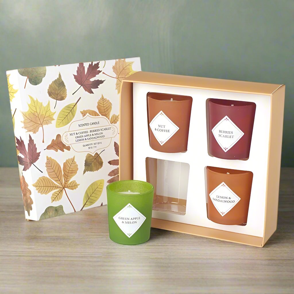 A gift box containing four scented candles with various color labels such as green, red, and brown, set against a background with autumn leaves.