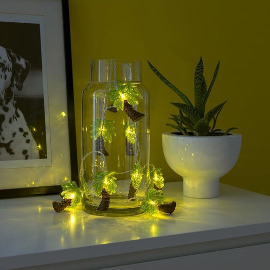 palm tree lights shown half in and half out a clear glass jar, the jar is on top of a white set of drawers with a green plant next to it in a white pot and half a picture frame leaning against the wall in the background