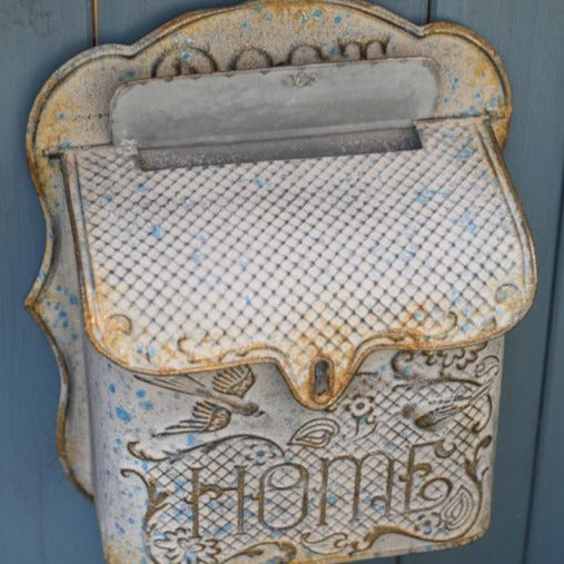 Rustic metal post box that says Home on the front. This post box is hanging on a painted blue wooden fence