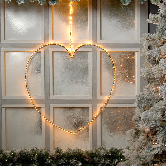 lit heart hanging in front of a snowy window with pinecone green garland and a snowy garland surrounding the window
