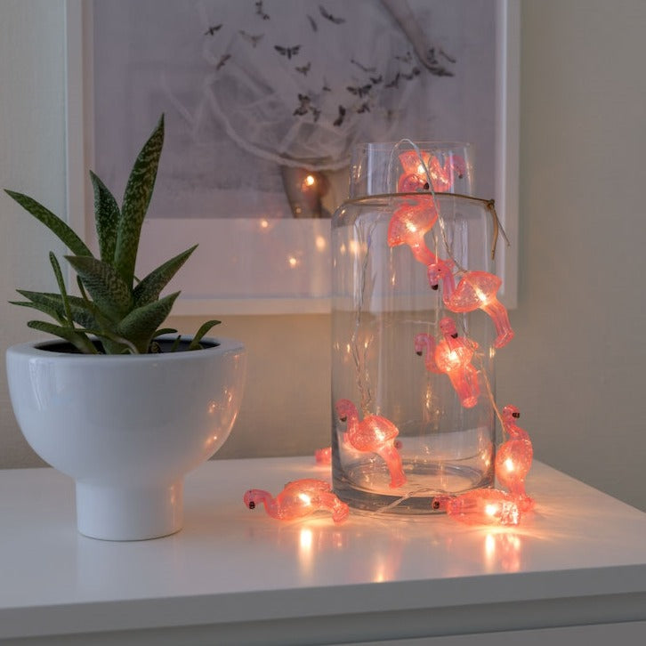 Konstsmide Flamingo Battery Lights half in half out a clear glass jar to show how they can be displayed. The glass jar is on top of a white set of drawers next to a green plant in a white pot and with a picture hanging on the wall in the background
