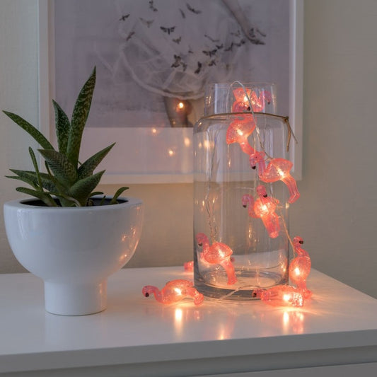 Konstsmide Flamingo Battery Lights half in half out a clear glass jar to show how they can be displayed. The glass jar is on top of a white set of drawers next to a green plant in a white pot and with a picture hanging on the  wall in the background