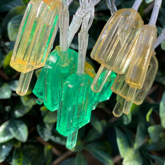 yellow and green ice lolly lights on a clear cable clumped together for the picture to show the lights off clearer. the background is a green leaf trellis