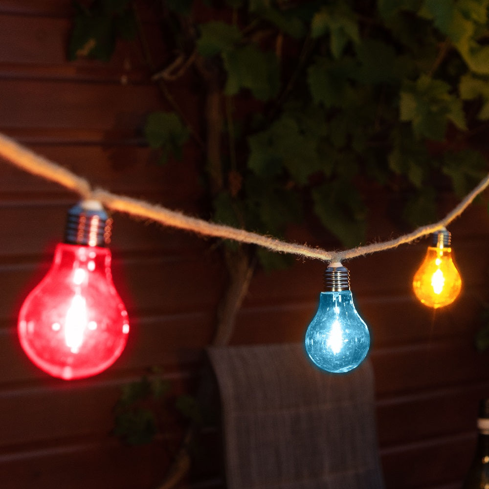 String of colorful light bulbs hanging against a wooden wall with plants.