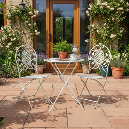 White metal garden chairs and table on a patio with flowers and plants around.