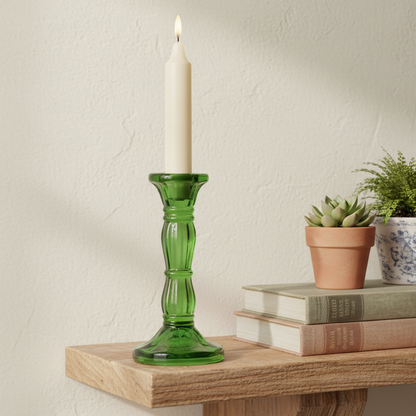 Green glass candlestick with a lit candle on a wooden surface with books and plants.