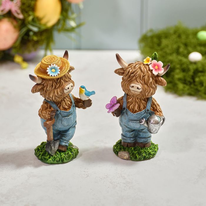 Two decorative Highland cow figurines with accessories on a light surface.
