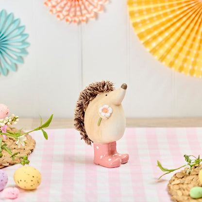 Decorative hedgehog figurine with pink boots and a flower, placed on a checkered surface with decorative fans in the background.
