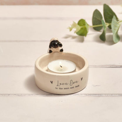 Ceramic tea-light holder with a candle and a small sheep figurine on a wooden surface with eucalyptus leaves in background