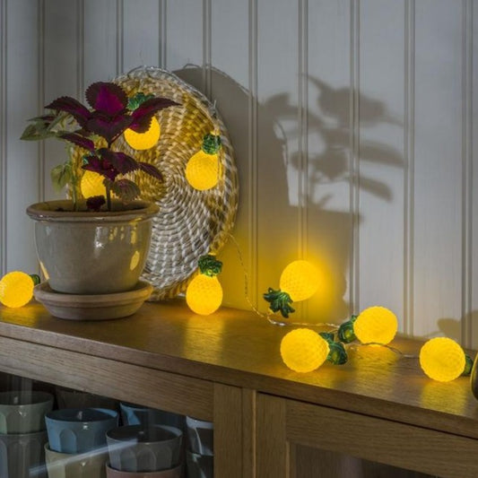 yellow pineapple LED lights shown half hanging on a wall and half lay on top of a dresser, with a purple plant in a pot in the picture in front of some of the lights.